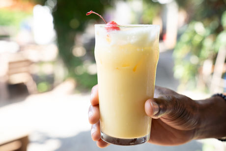 Hand holding a tall glass of creamy yellow drink with a cherry on top.