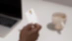 Hand holding a white credit card over a table with a laptop and a speckled mug. Neutral tones, minimalist office setting.