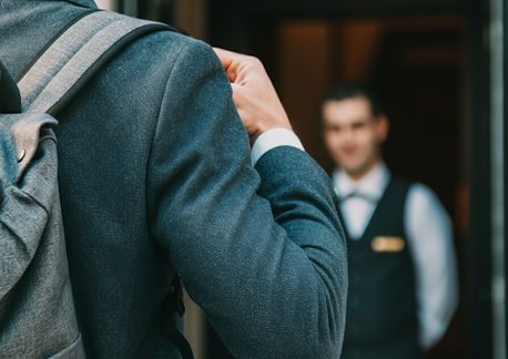 Person with backpack entering, greeted by smiling concierge at hotel entrance.