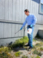 Man in a blue checkered shirt and jeans sprays weeds by a gray wooden house using a tank sprayer, wearing blue gloves, in a sunny setting.