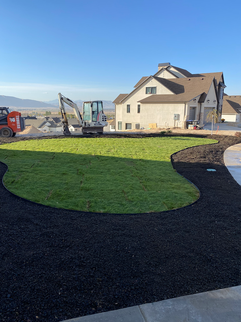 A construction site with a small excavator and pickup truck on a dirt road near a house. Fresh grass is laid on dark soil under a clear sky.