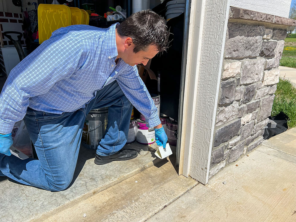 Man in blue plaid shirt kneels in garage near stone wall, holding pest control card in gloved hand. Bright sunlight. Cleaning supplies nearby.