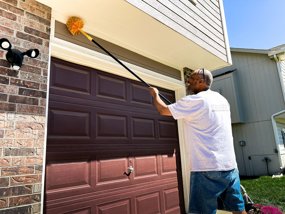Man cleaning a garage door frame with a long brush on a sunny day. He wears a white shirt and shorts. The garage is maroon.
