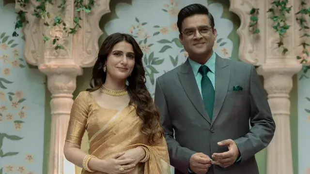 A woman in a gold saree and a man in a gray suit with a teal tie stand smiling in an ornate setting decorated with vines and arches.