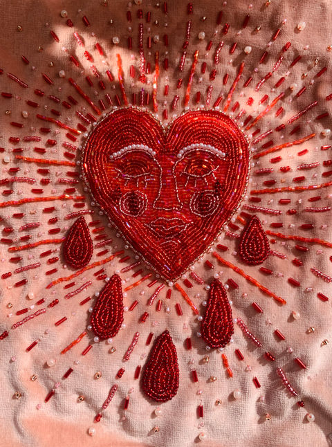 photo of beaded red heart on pink velvet fabric. the heart has a sleepy face and red droplets surround the heart. 