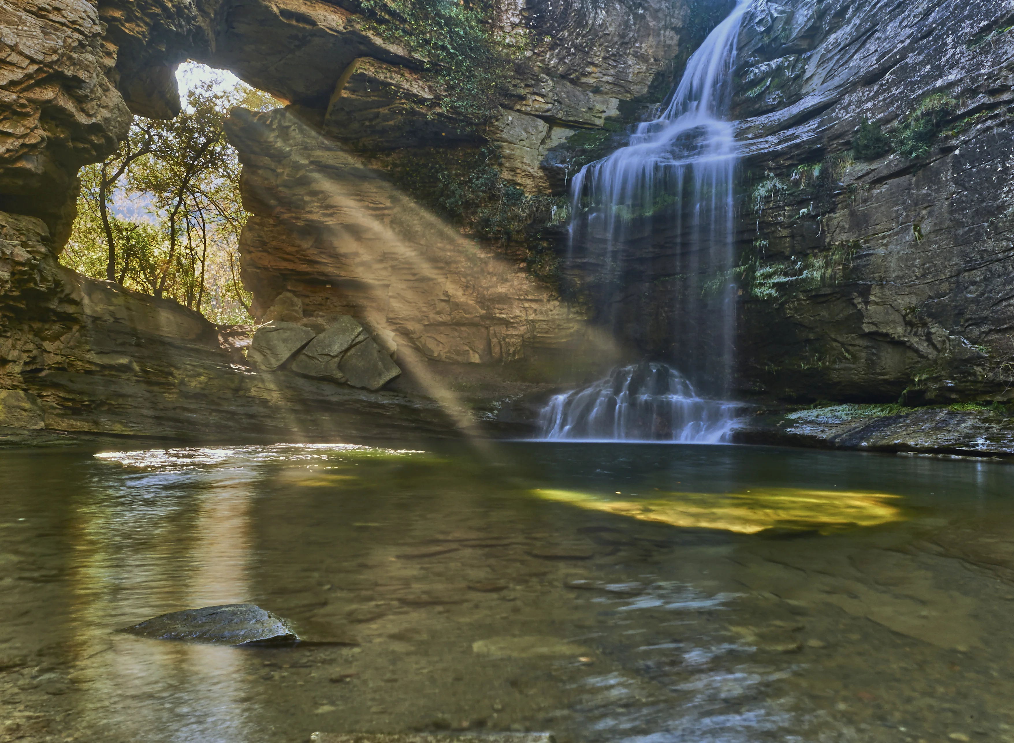 A waterfall next to a natural opening in the rock and sunlights streaming through it