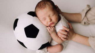 Newborn resting on big brother's soccer ball with brother's hands