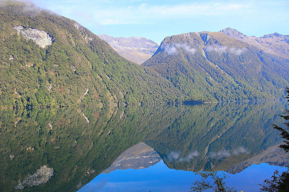 Montagnes verdoyantes se reflétant dans un lac calme, ciel bleu et quelques nuages, paysage paisible sans texte visible.