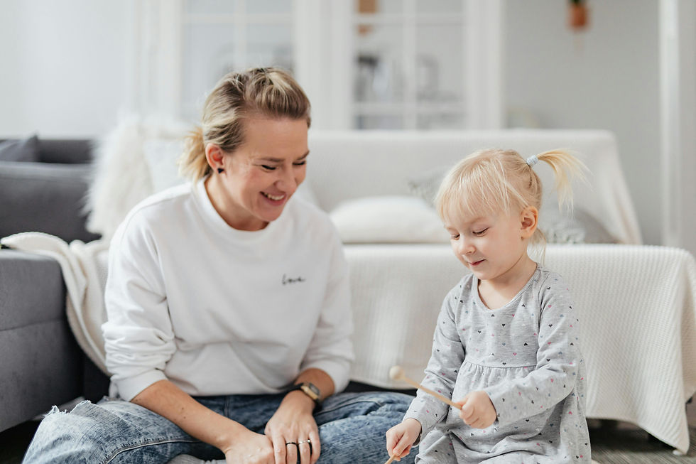 Adult and toddler playing happily on the floor in a cozy living room. Both are smiling. Child holds a stick; adult wears a white "love" top.