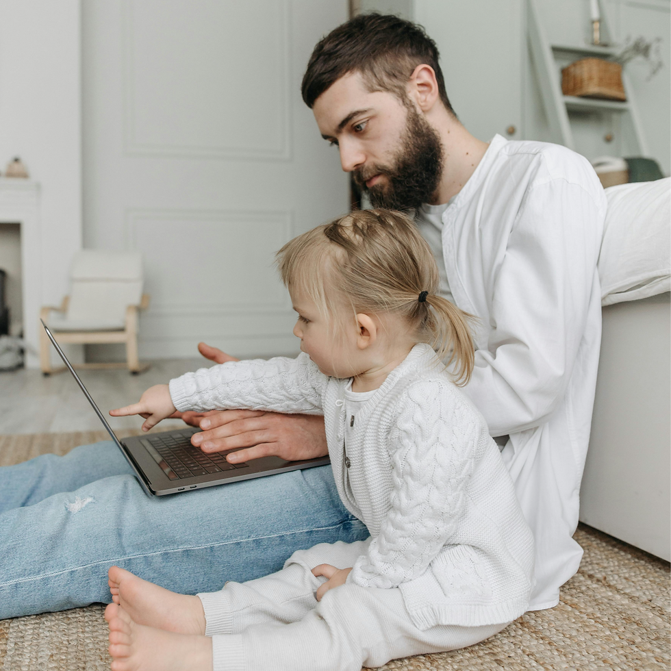 Adult and child sitting on the floor. Child points at a laptop screen; adult types. Both in white sweaters. Cozy room with light decor.