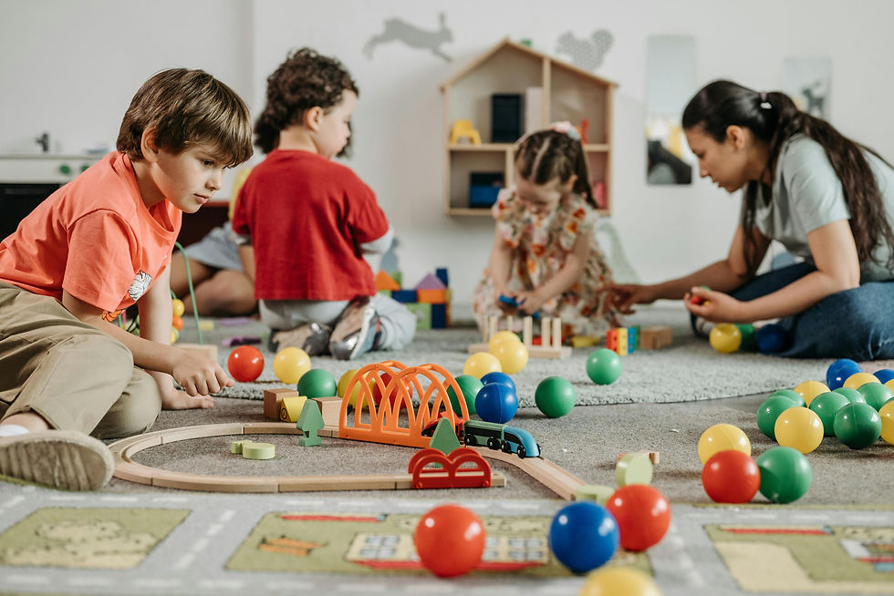 Children and a woman playing with colorful balls and toy blocks on a carpet in a playroom. A dollhouse and wall decorations in the background.