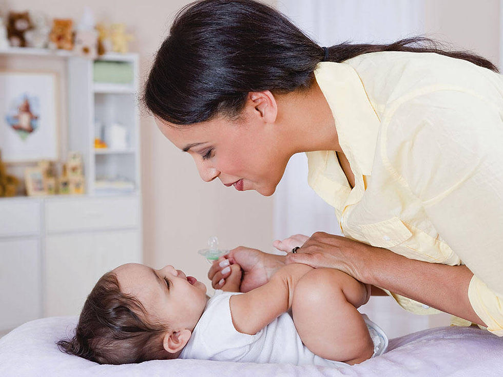Woman in a yellow shirt smiling at a baby on a bed, holding a pacifier. Cozy room with soft colors and toys in the background.