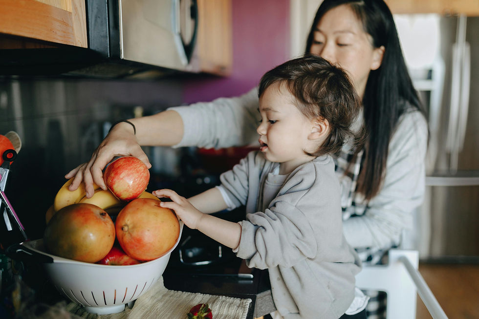 Child reaches for fruit in a colander while a woman holds an apple. Kitchen setting with purple and wooden tones. Cozy and focused mood.
