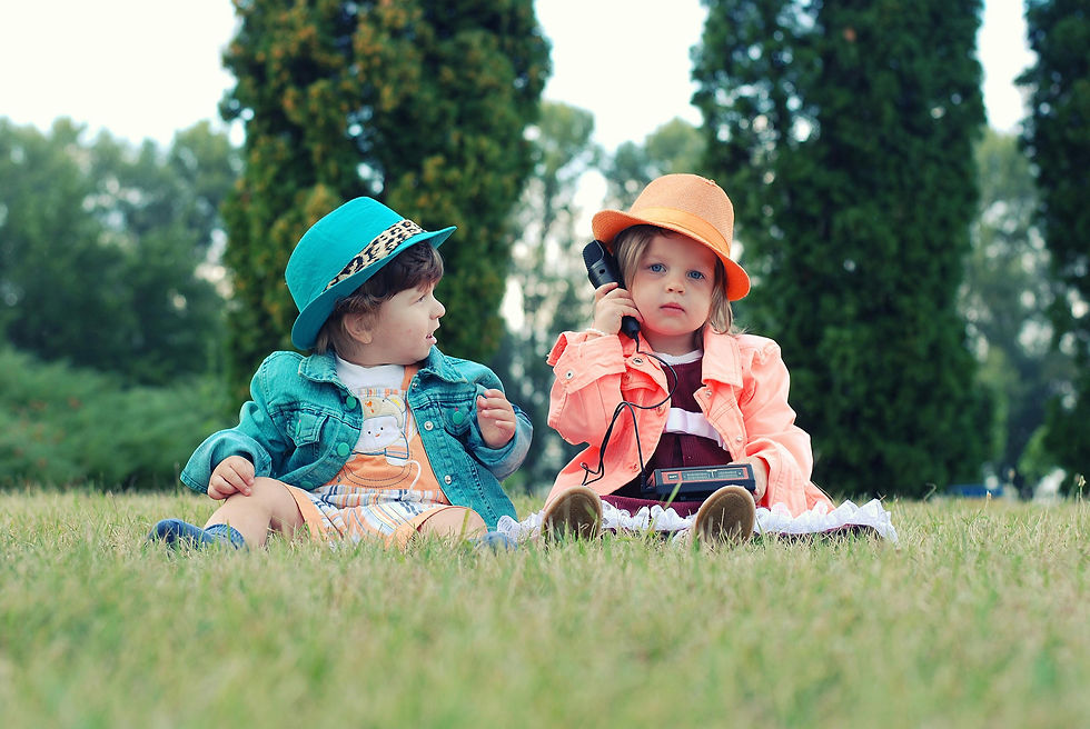 Two children in colorful jackets and hats sit on grass. One child holds a vintage phone, creating a playful mood in a park setting.
