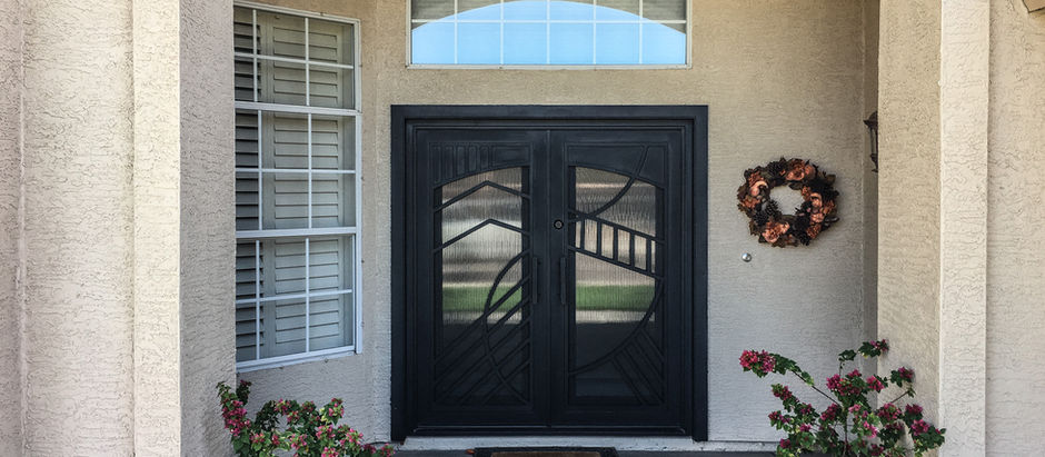 A black double custom iron entry door, a wreath, potted plants, and windows. Sunlit exterior with shadows on the walkway.