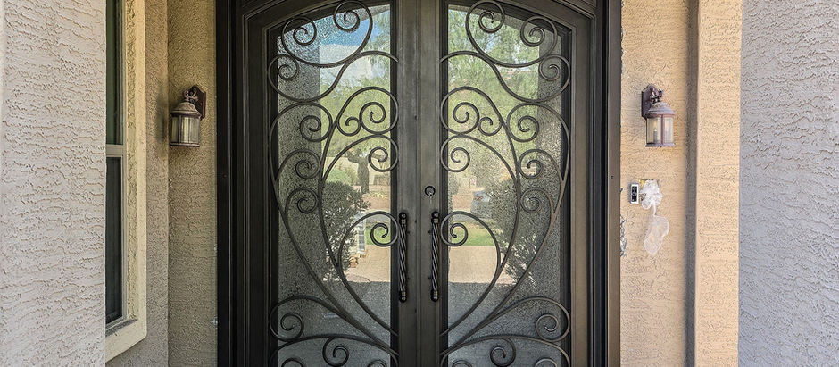 Elegant double iron entry doors with ornate black iron scrollwork, set in a tan archway. Brick path leads to the entrance. Sunlit, inviting ambiance.
