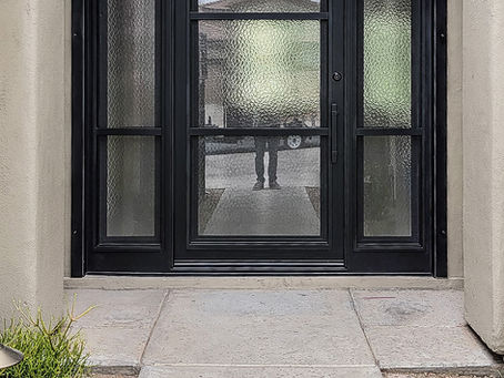 Black custom iron entry door with a patterned glass. Tiled path, and plants surround the entrance. Blue sky above.
