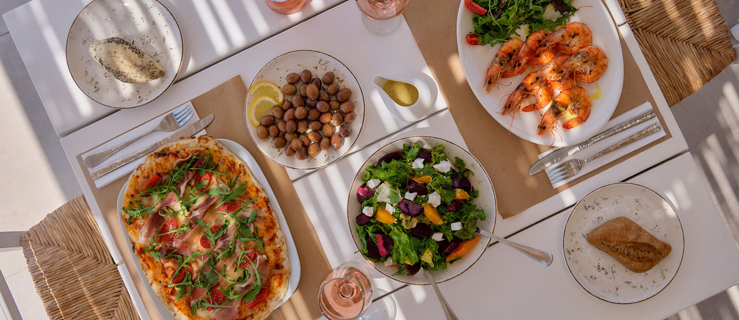 Overhead view of a lunch table at The Bird Café — prosciutto pizza, grilled prawns, beetroot and feta salad, olives and rosé wine