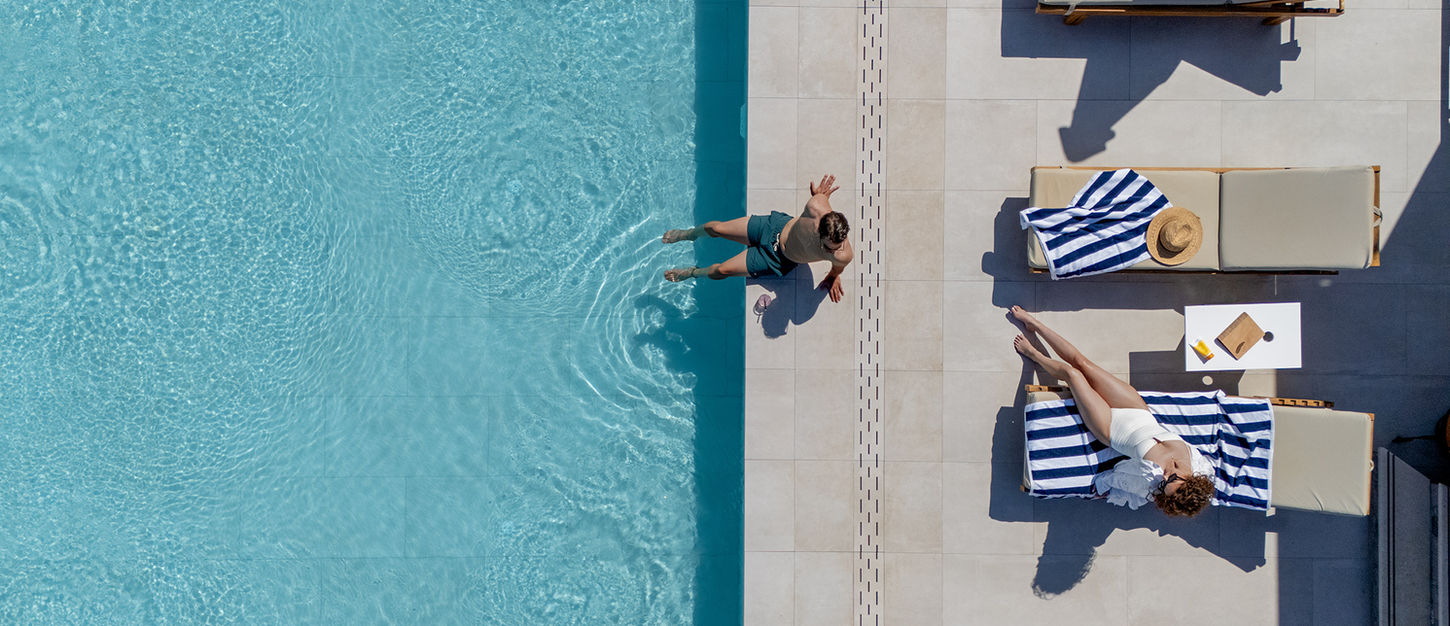 Aerial view of a man jumping into the infinity pool at Little Bird Lesvos while a woman sunbathes on a striped towel on the pool deck