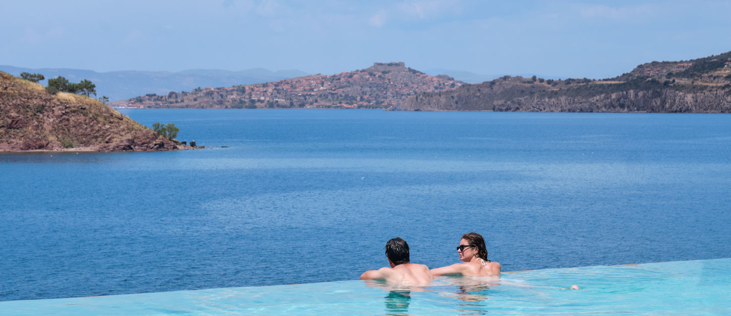 Couple in the infinity pool at Little Bird Lesvos looking out across the Aegean Sea towards St George's Island and Molyvos with its medieval castle