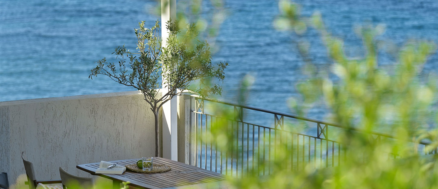 Villa terrace at Little Bird Lesvos with a timber table, book and drink, a small olive tree and the Aegean Sea beyond lush green foliage