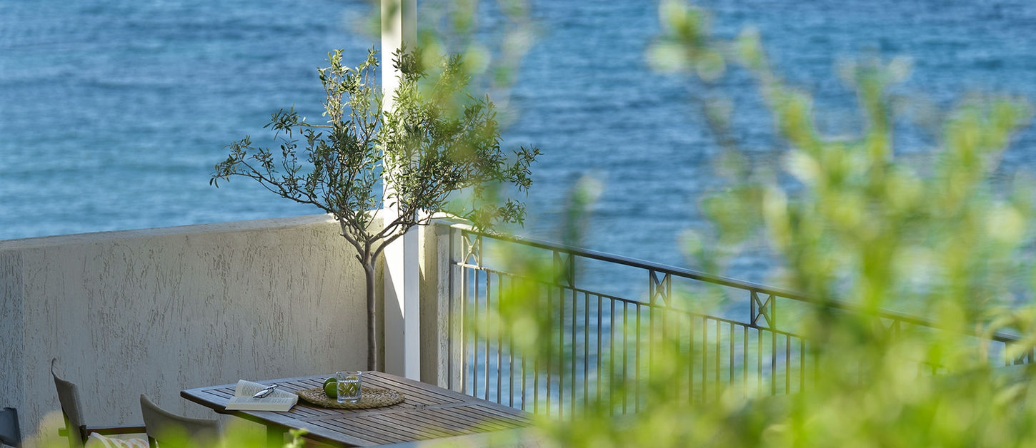 Villa terrace at Little Bird Lesvos with a timber table, book and drink, a small olive tree and the Aegean Sea beyond lush green foliage