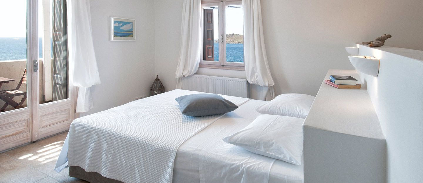 Double bedroom at Little Bird Lesvos with white linen, open French doors to a sea-view balcony and a small window framing St George's Island