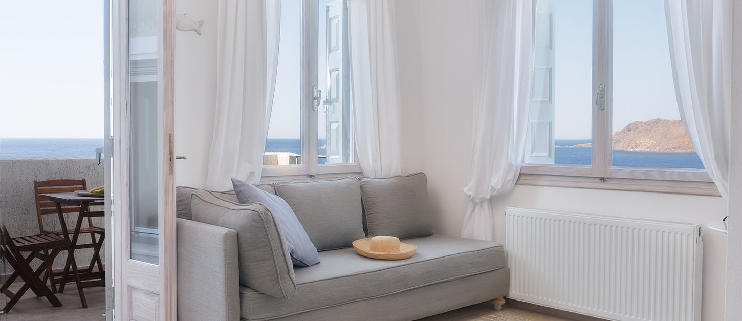 Light-filled villa living room at Little Bird Lesvos with a grey sofa, sun hat on the cushion, open French doors to a balcony and sea views