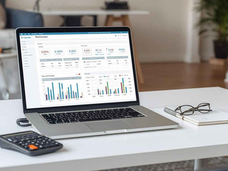 a laptop displaying financial charts and data sits on a white desk, accompanied by glasses, a notebook, and a calculator, suggesting a workspace setting for virtual bookkeeping.