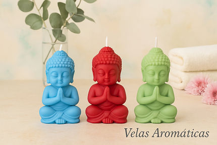 Velas aromáticas en forma de Buda en colores azul, rojo y verde, con manos en posición de oración, sobre superficie clara con fondo de toallas blancas, ramas de eucalipto y decoración floral, estilo spa.