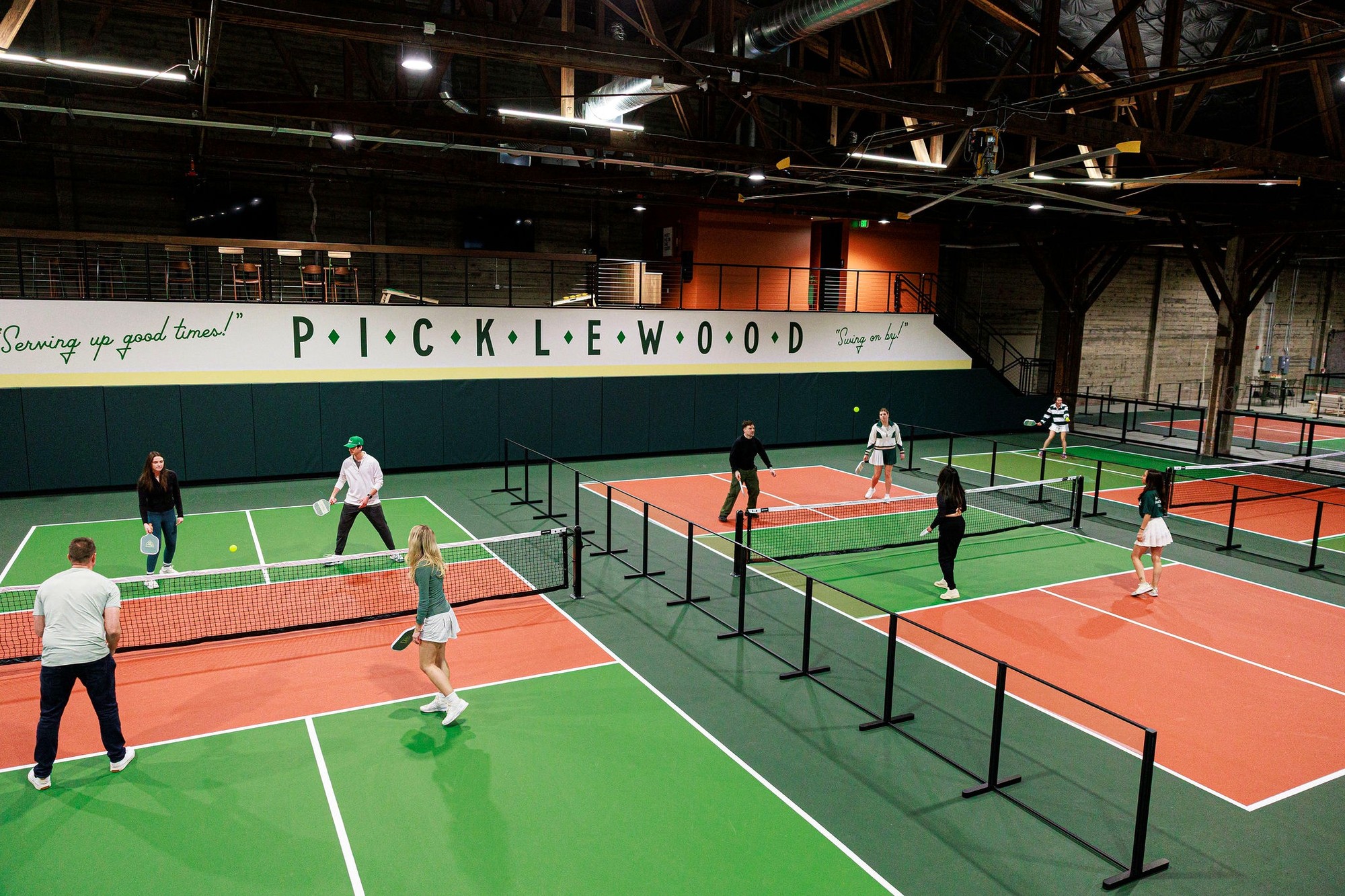 Players mid-rally across three of Picklewood's indoor courts in SoDo, paddles up, under the club's painted wall sign.