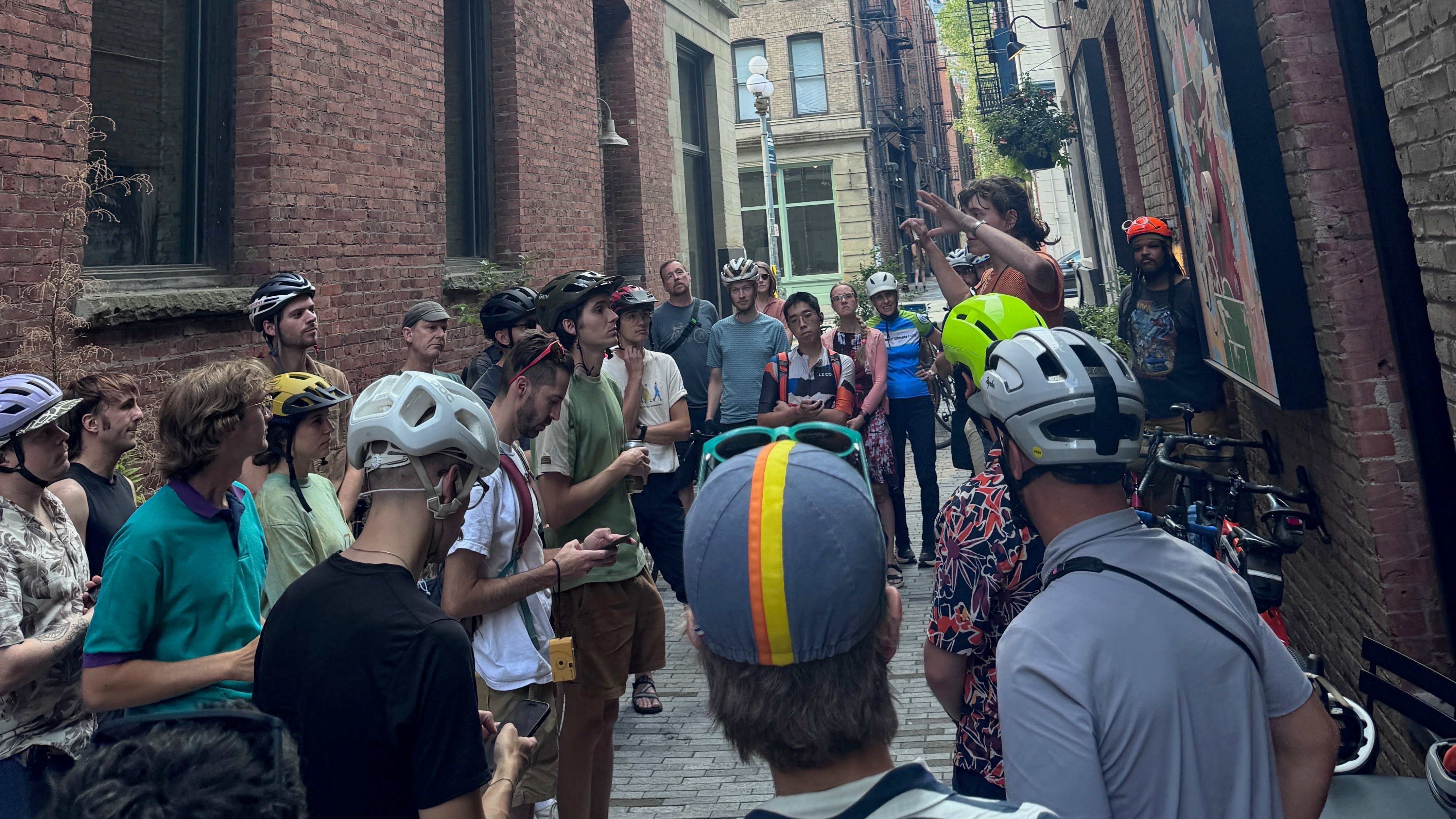 About twenty-five riders in helmets and caps packed into RailSpur Alley between brick walls, listening to a ride leader before a Sound Break ride. The shop's own photo (IMG_5048), cropped 16:9; owner approved 2026-10-14.