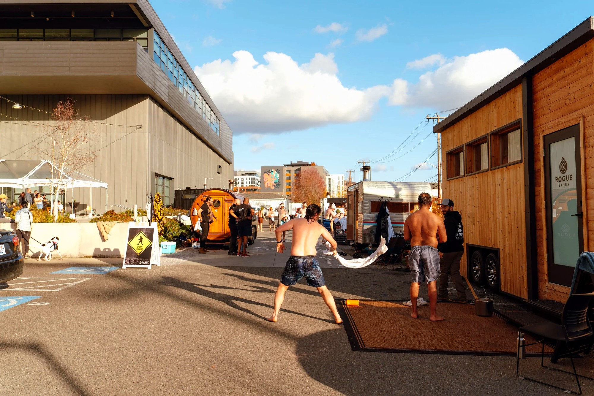 Bright sunny day in the alley between sauna stations: a man mid-motion swings a towel toward another man standing barefoot beside the 'Rogue Sauna' cabin, a leashed dog crosses in the foreground, a yellow 'SAUNA CROSSING' sign stands on the pavement, the museum building visible behind.