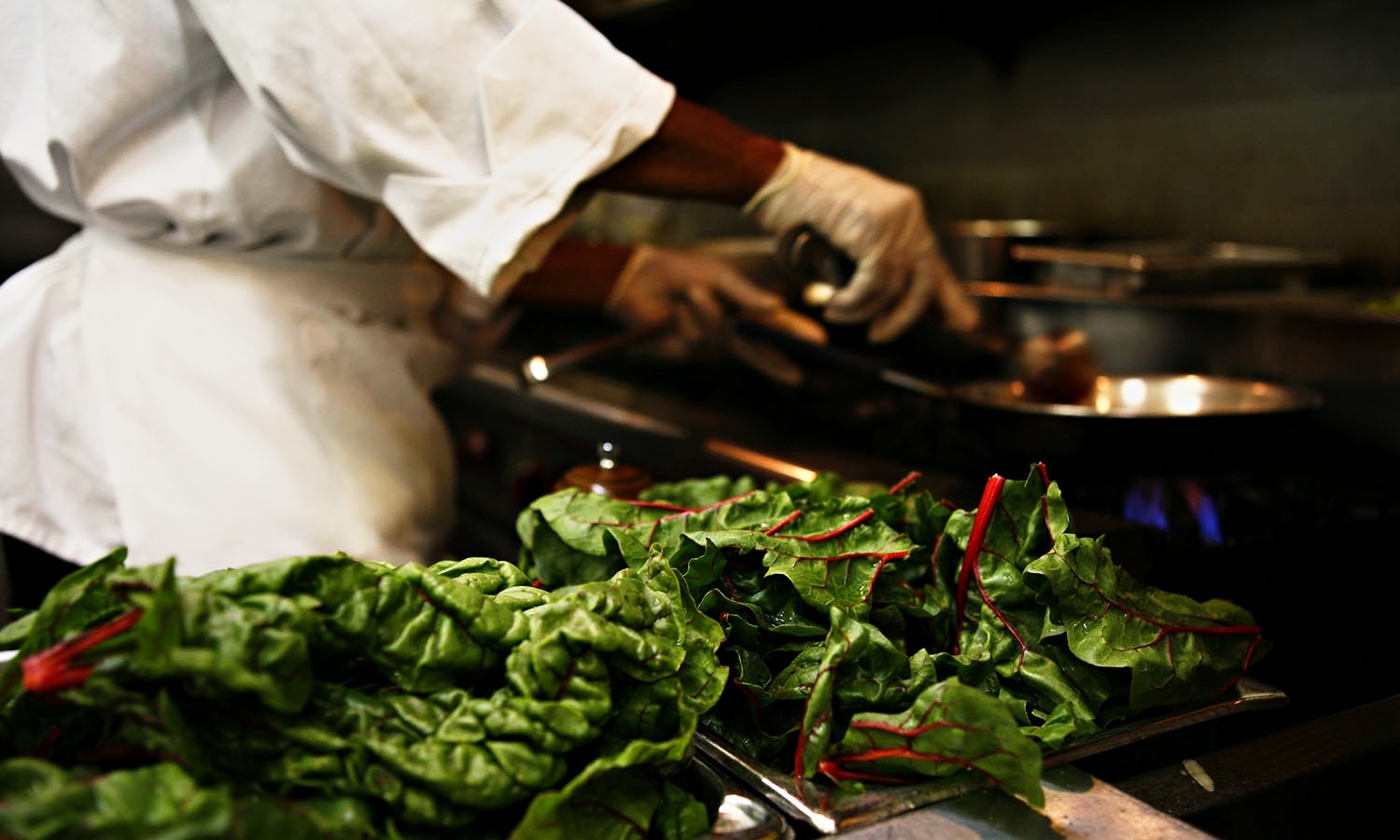 A FareStart cook in kitchen whites works a pan on a lit gas range behind a tray of freshly cut rainbow chard.