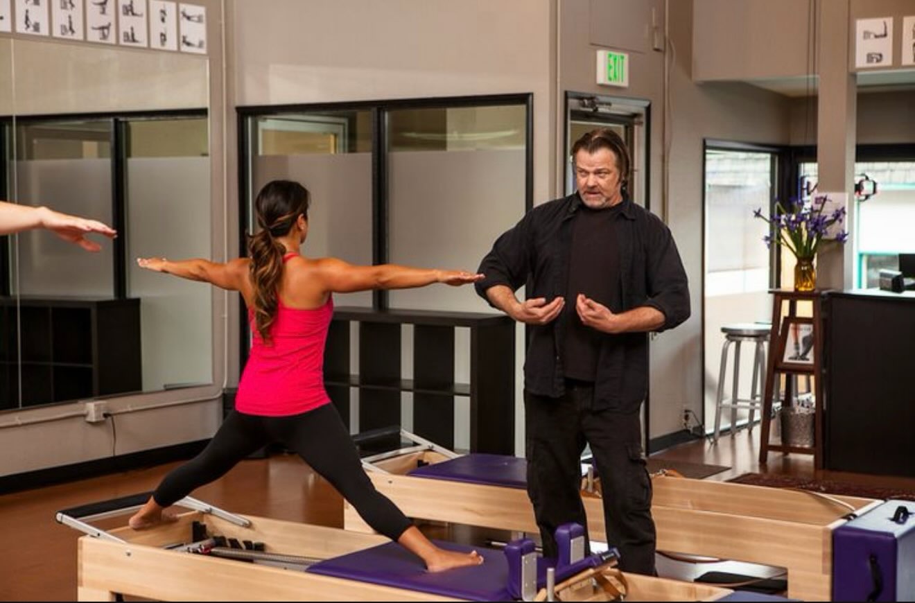 Tom Wiseley, standing, coaching a woman in a pink top holding a lunge with arms out on a Pilates reformer, bright studio with mirrors behind