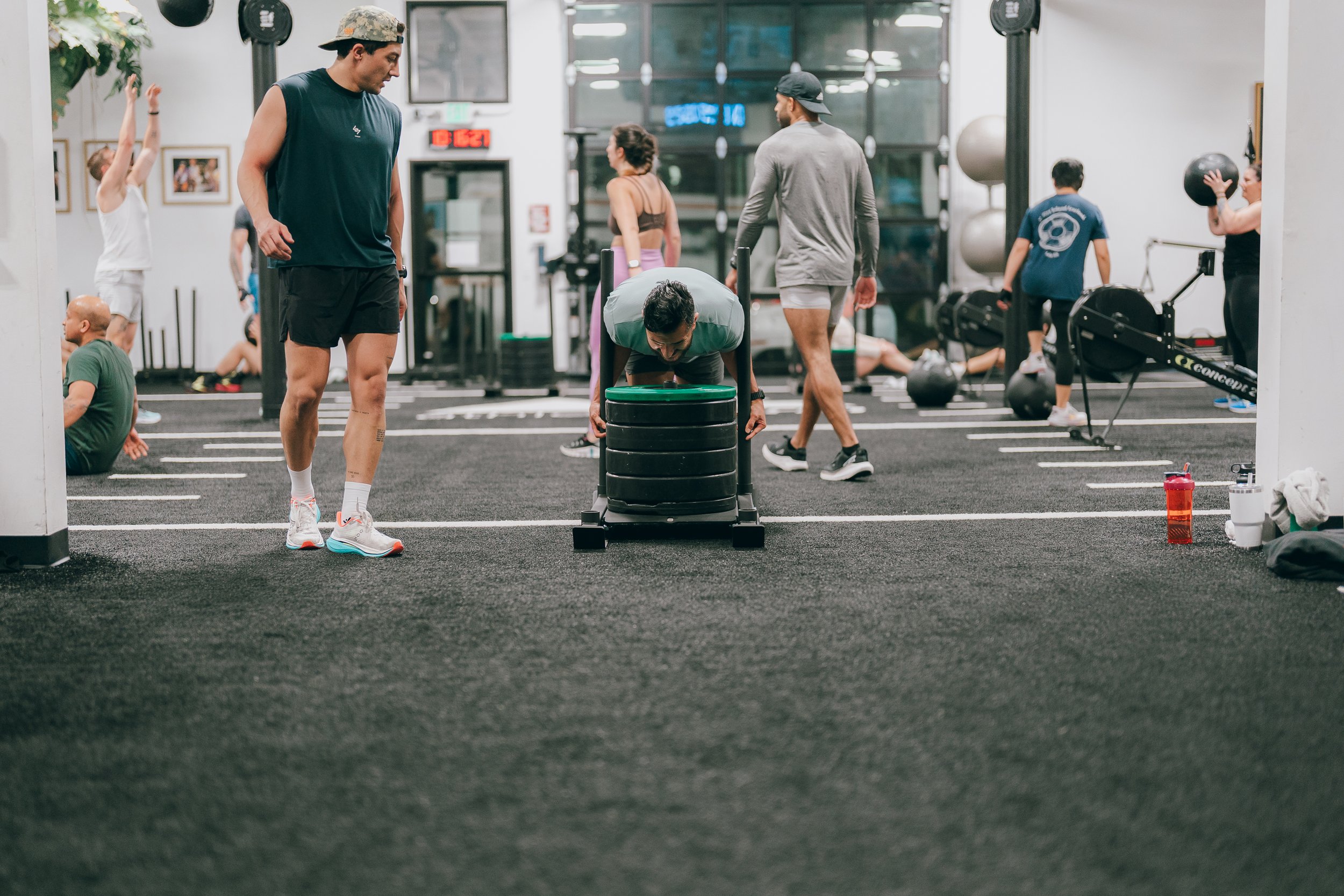 A member driving a sled push mid-rep on Athletic Form's SoDo floor, a coach watching, others on wall balls and a rower behind.