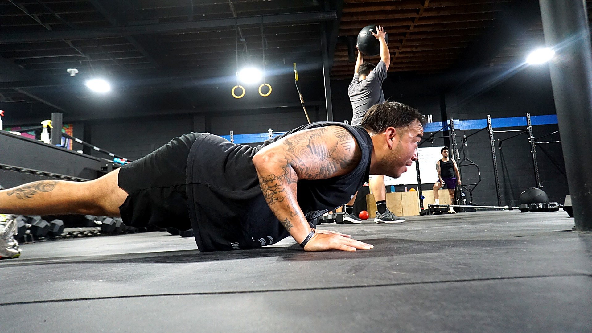 A member mid-burpee in the foreground and another mid-wall-ball throw behind him, in Root Strength's black-painted Georgetown unit, rings overhead.