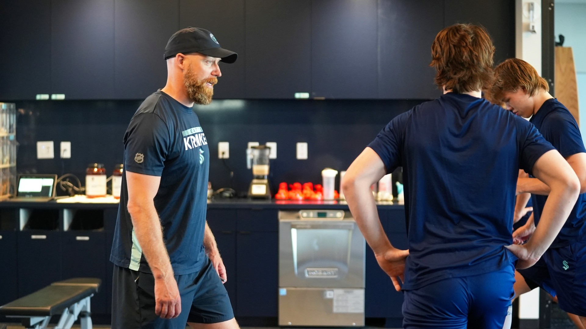 A man in a black Seattle Kraken staff t-shirt and cap — Kraken logo on the chest — stands and speaks with two tall young players in navy Kraken-branded shorts and shirts in what appears to be the facility's nutrition kitchen or recovery area. Dark navy cabinetry, a commercial dishwasher, blenders, and red recovery drink bottles are visible on the counter behind them. The staff member faces the players directly; the players are turned away from camera. Everyone is big in the frame.