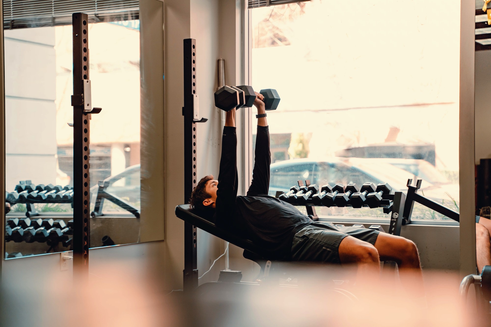 A man lying on a flat bench inside a gym, pressing two hex dumbbells overhead at full extension with both arms. He wears a black long-sleeve shirt and dark shorts. Behind him: a power rack, a full dumbbell rack, and large front windows looking out to a street with a parked car and building visible. Bright natural window light floods the room. The setting is clearly a gym interior.