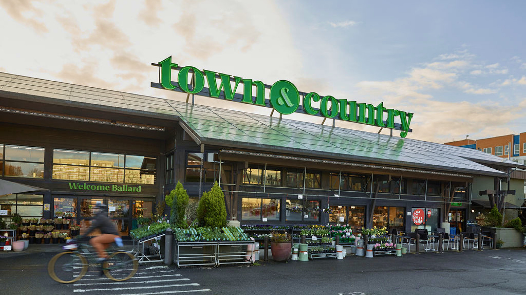 The Town and Country Markets storefront in Ballard at dusk, a large green town and country roof sign and a Welcome Ballard banner both lit, flower and plant displays along the front, a cyclist riding past.