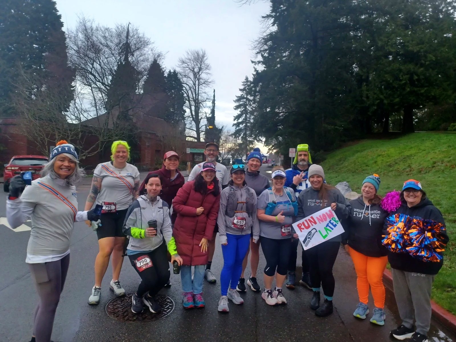About thirteen to fourteen people posed as a group on a wet sidewalk/street corner on a grey, overcast day. All are dressed in running gear — tights, running tops, hats, gloves, race bibs visible on some. One person holds a handwritten sign reading 'RUN NOW LATER'. At least one person wears a shirt with 'West Seattle Runner' text visible. One person holds blue and orange pom-poms. Bare winter trees and a brick building (appears to be a church) are in the background. The group is looking at the camera, smiling.