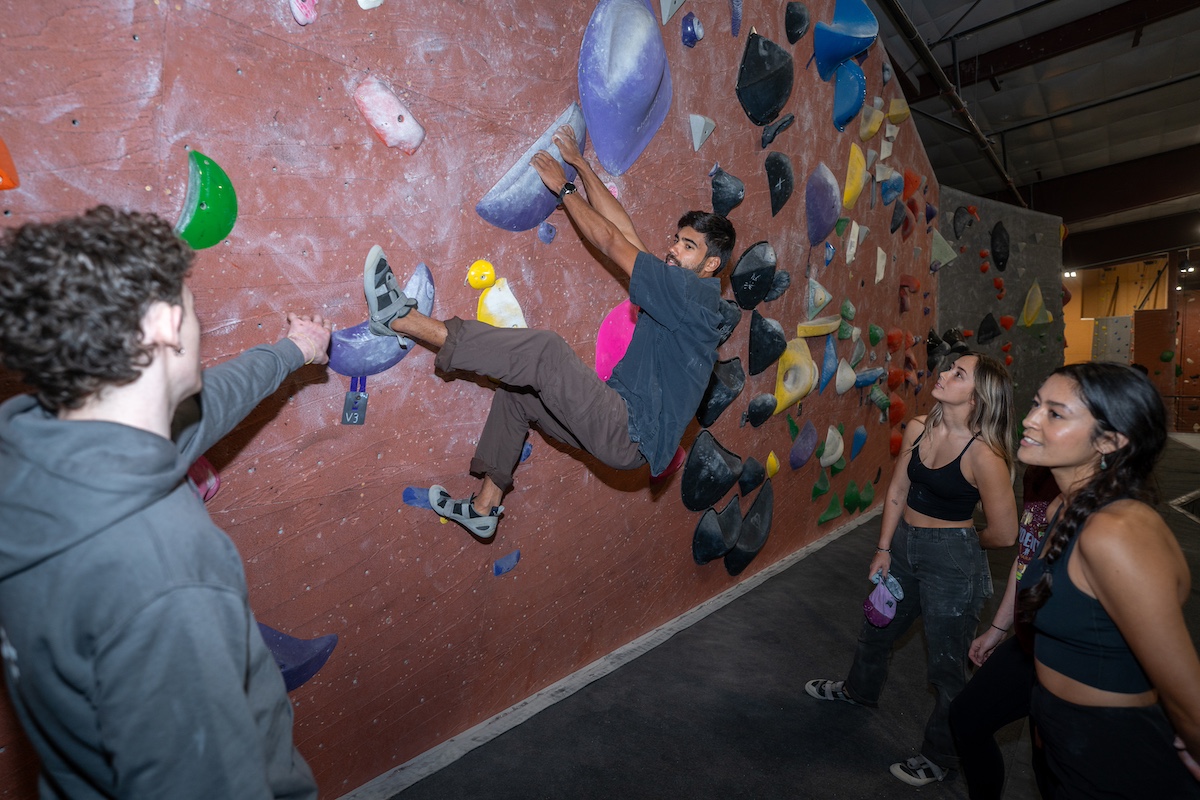A climber mid-move across a bouldering wall at Vertical World, spotters and onlookers beside him.