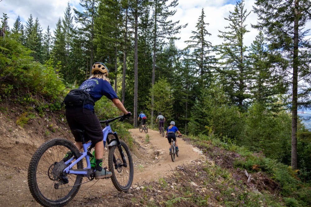 Four mountain bikers riding in a line down a dirt singletrack trail cut into a forested hillside. The lead rider is large in the frame — helmet, backpack, blue jersey, light-blue full-suspension bike — with three riders behind receding into the distance. Conifer forest flanks both sides; a glimpse of water or a valley is visible through the trees at the right edge. All riders wear helmets and MTB gear. Active descent, bodies weighted, engaged with the trail.