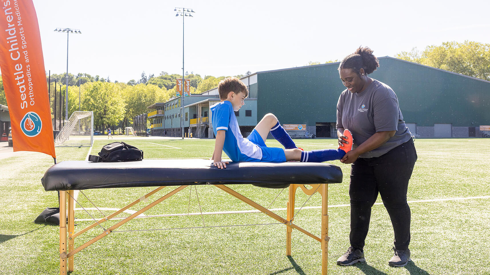 A Seattle Children's athletic trainer examining a boy's ankle on a portable treatment table at the edge of a turf field, an orange Seattle Children's Orthopedics and Sports Medicine banner beside them.