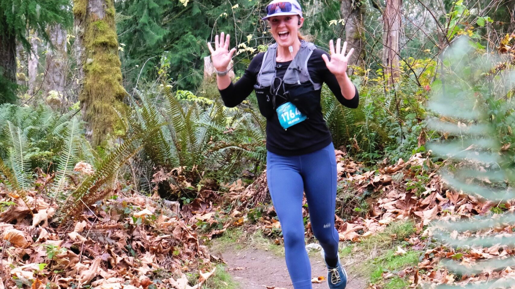 A single woman runner on a forest trail, bib number 1764 visible, arms spread wide with hands raised and a huge grin toward the camera, mid-stride on a leaf-covered dirt path. Mossy ferns, autumn leaves, and tall conifers surround her. She is big in the frame and clearly in motion.