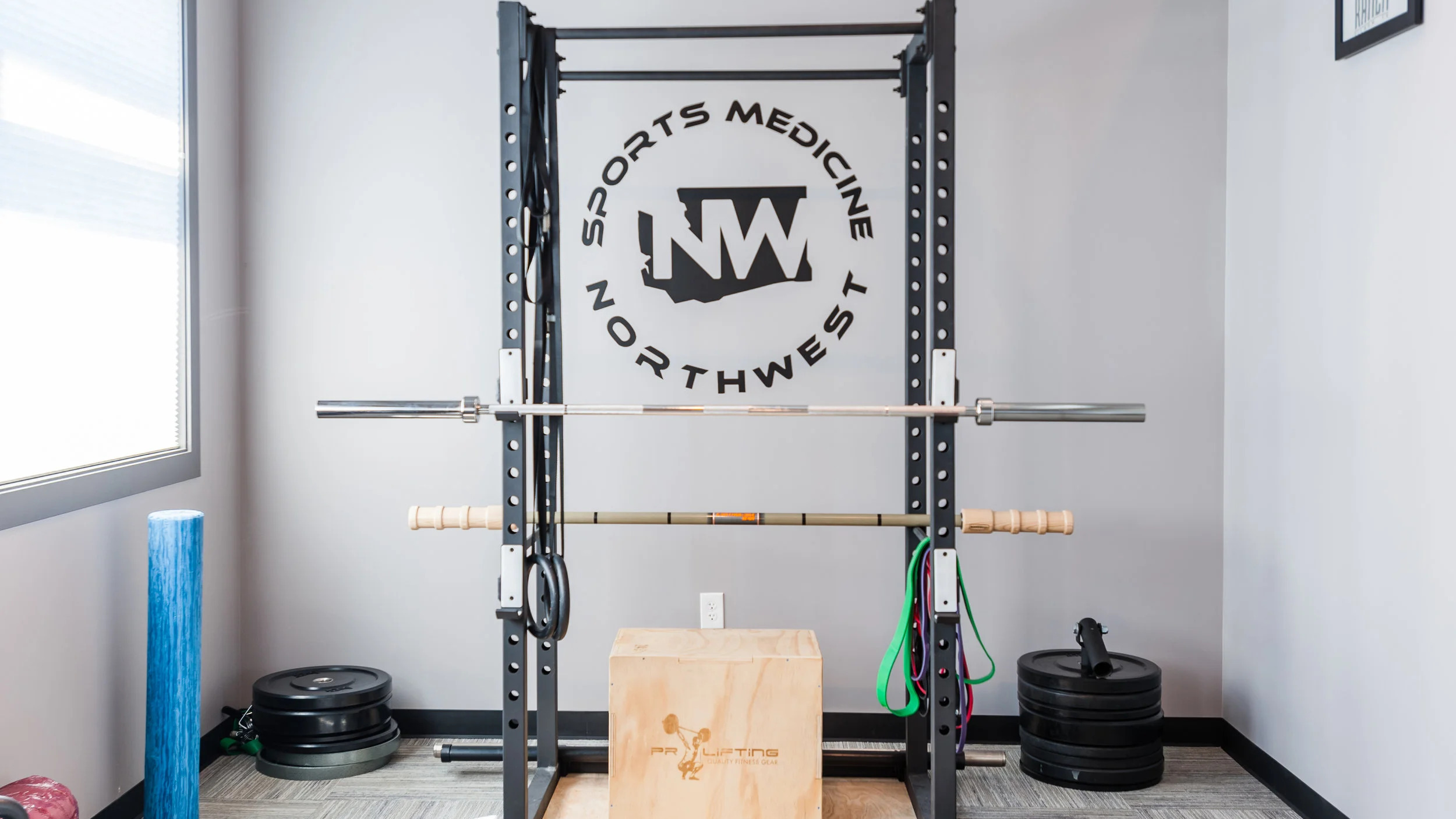 A power rack in a training room with a large Sports Medicine Northwest logo, a circle around a Washington state outline, mounted on the wall behind it. A loaded barbell, a wooden plyo box, resistance bands and weight plates.