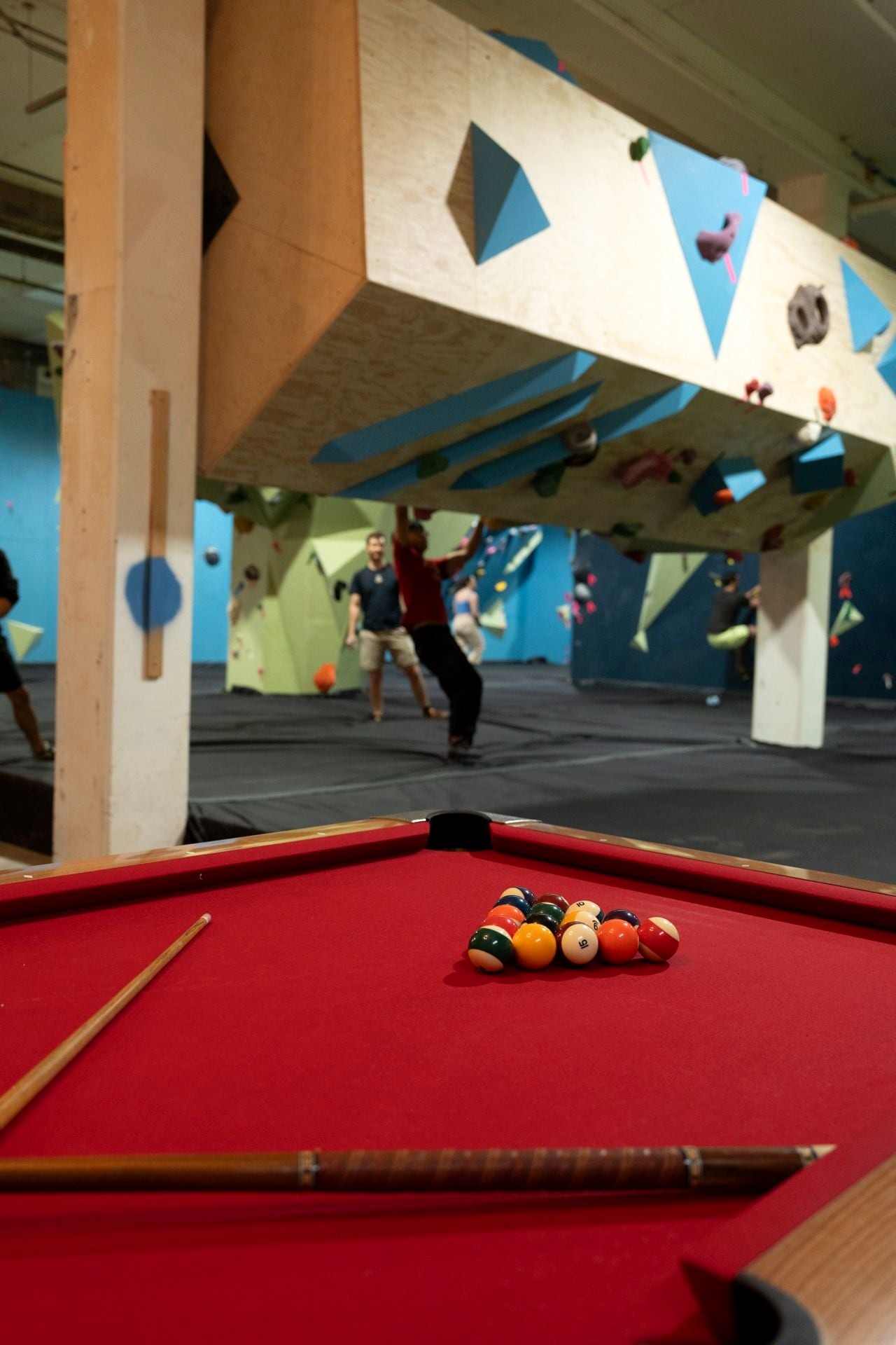 Foreground: a red-felt pool table with balls racked. Directly overhead, seen from below, the gym's 'rolling pin' rotating climbing feature — a huge plywood-and-blue-panel cylinder studded with holds, suspended on posts. Beyond it, two members are mid-boulder on green and blue walls under it, one with arms raised on a hold, one climbing past a corner arete.