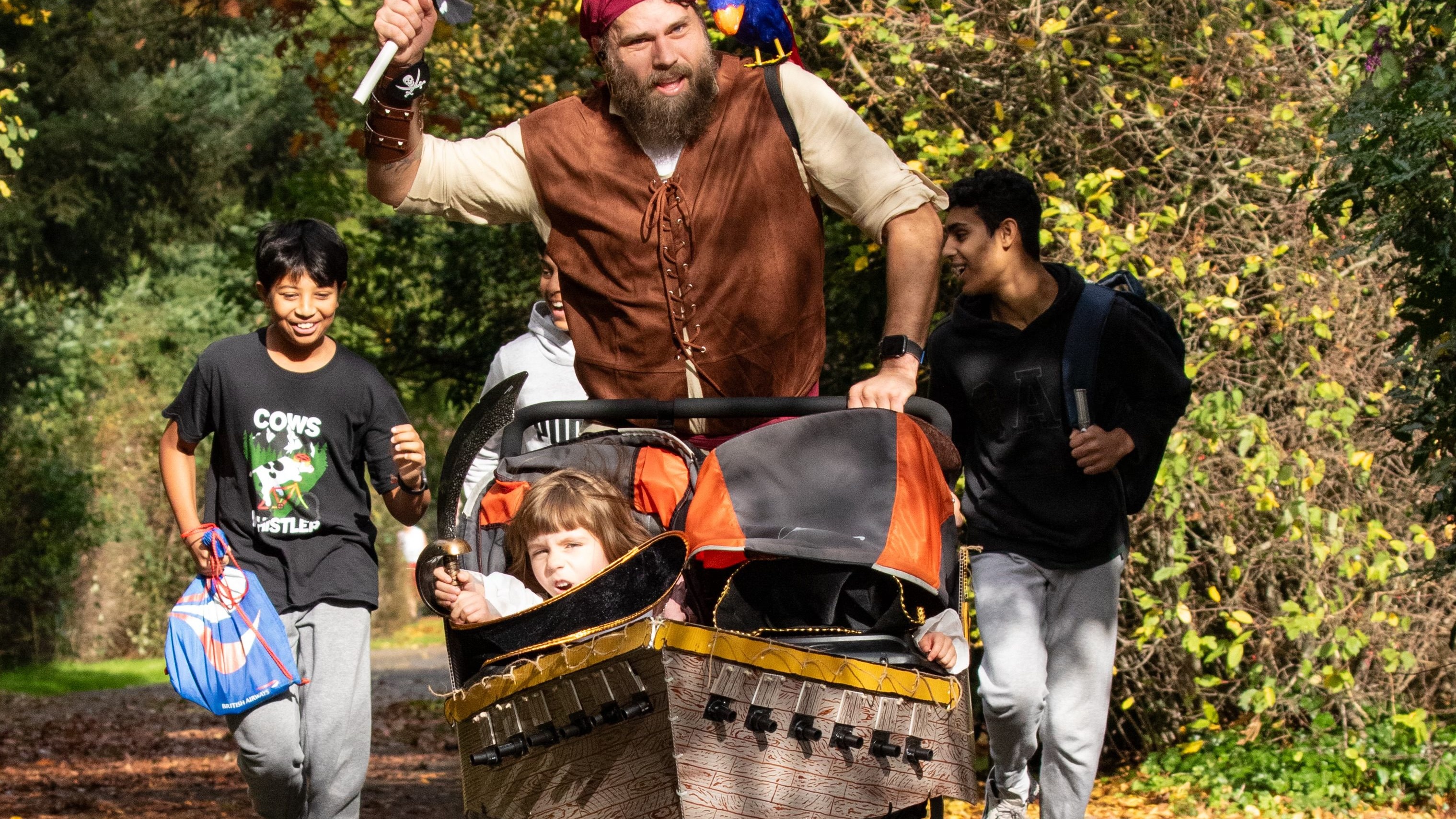 A group of people walking along a leafy paved trail in bright autumn light. In the centre a bearded man in a full pirate costume (brown leather vest, bandana, small parrot on shoulder) pushes a baby stroller decorated to look like a pirate ship/treasure chest, with a toddler inside. Three other people walk alongside — a young man on the left in a 'Cows & Hustlers' T-shirt, two others to the right — all smiling or laughing. Autumn trees with yellow leaves line the trail. Nobody is running; all are walking. No race bibs visible on the non-pirate participants.
