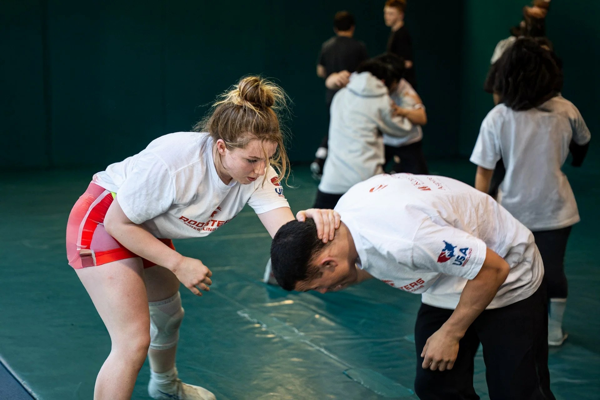 A wrestling practice session on a teal mat inside a gym with teal-painted walls. In the foreground a young woman in a 'Roosters Wrestling' white t-shirt and red wrestling shorts/knee pad has her hand on the head of a male partner who is bent over; both are in active drill stance. Several more wrestler pairs work in the background.