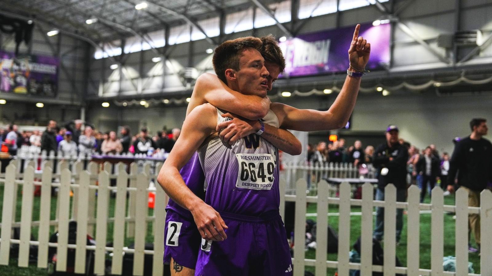 Two UW male runners in purple Washington Huskies kit embracing and celebrating at the finish area of Dempsey Indoor. The runner facing the camera (bib 6649, 'WASHINGTON / DEMPSEY INDOORS') hugs a teammate from behind; one arm is raised with index finger pointing upward in a #1 gesture. A large crowd fills the bleachers behind them and the white picket track fence is visible. The bib text reads 'WASHINGTON / DEMPSEY INDOORS' confirming the venue.